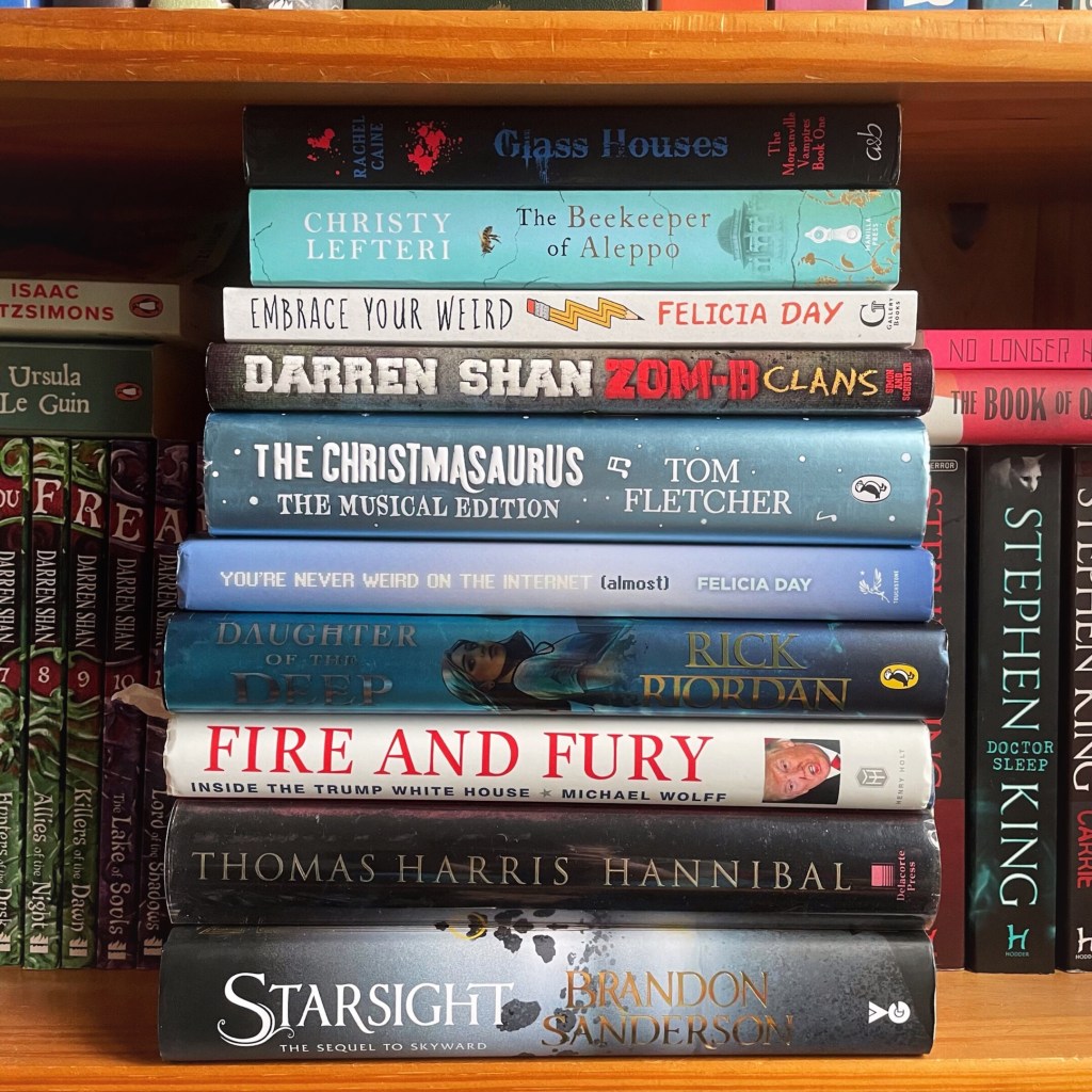 Stack of books on a book shelf. Overlayed on the image is a red box with the words FAQ. The books are Starsight by Brandon Sanderson, Hannibal by Thomas Harris. Fire and Fury by Michael Wolfe, Daughter of the deep by Rick Riordan, You're never weird on the internet (Almost) by Felicia Day, The christmasaurus by Tom Fletcher, Zom-B Clans by Darren Shan, Embrace your weird by Felicia Day, The beekeeper of Aleppo by Christy Lefteri and Glass houses by Rachel Caine. 